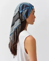 Pañuelo cuadrado de seda con estampado gráfico y geométrico en tonos azules, negros y blancos, adornado con flecos en los bordes.