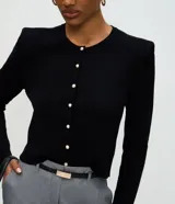 Cardigan de punto color blanco, de corte regular y ajuste al cuerpo. Presenta cuello redondo, mangas largas, cierre frontal con botones delicados y hombreras estructuradas que aportan volumen a la silueta.