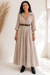 Vestido largo beige de seda, con cuello camisero, cierre frontal con botones y mangas largas remangables. Incluye un cinto marrón trenzado con hebilla dorada.