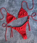 Conjunto de bikini rojo con corpiño triangular con tirantes finos y detalle de aro metálico con forma de corazón en el centro. La bombacha es tipo colaless con tiras finas ajustables en los laterales con detalle de aros metálicos.