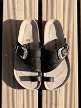 Sandalia de dedo color negro, confeccionada en cuero grabado, con plantilla anatómica, suela de corcho y hebilla ajustable con detalles metálicos estilo cowboy.