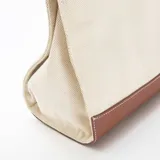 Bolso tipo tote de estructura rígida, confeccionado en lona color beige con detalles de cuero sintético en color marrón en la base, las asas y los bordes superiores.