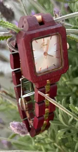 Reloj de pulsera con caja cuadrada y correa de eslabones de madera color borgoña. El dial es plateado con agujas y detalles dorados.
