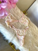 Conjunto de lencería compuesto por un bralette y una tanga, confeccionados en encaje bitono rosa y champagne con diseño floral. El bralette presenta cierre frontal con broche de gancho y breteles regulables. La tanga es de estilo colaless con laterales regulables.