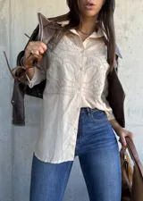 Camisa beige de corte oversize, con cuello clásico y cierre frontal de botones. Presenta un diseño bordado detallado en la parte superior delantera, con mangas largas y un estilo versátil.