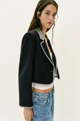 Blazer corta de color negro con cuello solapa, manga larga y hombreras. Presenta detalles combinados con estampado de cuadro vichy en el cuello, los bordes de la solapa, el ruedo y los puños. Cuenta con cierre frontal de un botón y falsos bolsillos delanteros de vivo.