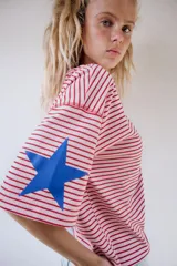 Camiseta oversize de rayas horizontales rojas y blancas, con cuello redondo y mangas cortas. Presenta un estampado de texto azul en el pecho y estrellas azules en las mangas.