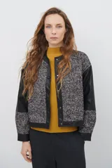Chaqueta de diseño combinado con cuerpo en tejido de lana con patrón de espiga y mangas de cuero sintético. Presenta cierre frontal con botones metálicos a presión, cuello redondo y bolsillos delanteros.