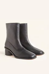 Bota de caña media confeccionada en cuero vacuno negro, con diseño minimalista y cierre trasero. Presenta un taco cilíndrico de madera tallada y punta cuadrada.