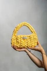 Cartera de mano tejida a crochet en hilo de algodón reciclado, color amarillo. Presenta un diseño compacto con manija integrada y cierre de botón imantado.