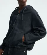 Campera tipo bomber confeccionada en tejido de jean, con cierre frontal de cremallera, capucha ajustable con cordones y bolsillos frontales. Presenta puños y cintura elásticos, ofreciendo un ajuste cómodo y un estilo urbano.