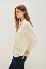 Camisa de algodón color crudo con diseño de encaje calado y detalles de plisados verticales en el frente. Presenta cuello clásico, cierre frontal de botones y mangas largas con puños delicados.