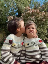 Sweater tejido a mano en lana merino, con cuello redondo y diseño con rayas horizontales en tonos crudo y marrón. Presenta bordados de animales y flores en la parte superior. Tiene puños y ruedo acanalados en color bordó.