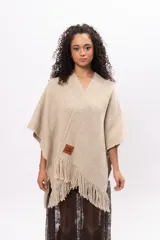 Ruana de tejido de punto en color arena, con diseño cruzado, escote en V y terminaciones con flecos en el ruedo. Incluye un parche de cuero con la marca Pura Lana en la parte frontal.