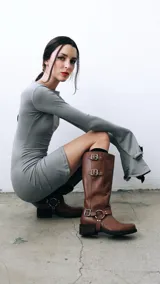 Bota de caña alta confeccionada en cuero color suela, con diseño de estilo western. Presenta tres hebillas metálicas en la parte superior de la caña y un estribo con argolla metálica en el tobillo. Cuenta con taco cuadrado de altura media.