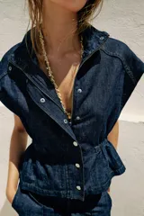 Chaleco de denim azul oscuro con cuello alto, manga sisa y cintura ajustable con elástico. Cierre frontal con botones a presión metálicos.