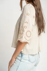 Blusa color arena de algodón peinado, con escote redondo, mangas cortas abullonadas con bordados suizos y detalles de alforcas en el frente.