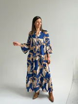 Vestido largo azul con estampado de hojas en tonos beige y blanco, escote en V con botones frontales, mangas largas amplias y diseño escalonado.