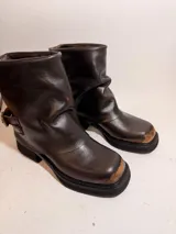 Bota de caña media confeccionada en cuero con efecto desgastado, presenta un diseño holgado en la parte superior y una tira con hebilla metálica ajustable en el tobillo. Cuenta con suela de plataforma y taco cuadrado robusto.