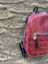 Mochila color bordó con textura tipo reptil en la parte superior y bolsillo frontal con diseño capitoneado en material charolado. Cuenta con cierre metálico y tiradores con flecos decorativos.