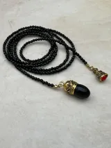 Collar largo de cuentas negras facetadas con terminaciones en dijes metálicos dorados, uno con piedra negra ovalada y otro con piedra roja pequeña.