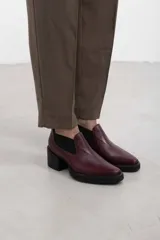 Botín asimétrico de estilo Chelsea color bordó, confeccionado en cuero vacuno, con paneles elásticos laterales negros, puntera cuadrada, suela de plataforma y tacón de bloque cuadrado.