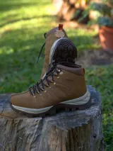 Botín de trekking color tostado con cordones, suela de goma dentada y refuerzos laterales. Presenta cuello acolchado y detalles metálicos en los ojales.