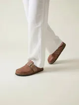 Zueco de cuero color marrón con diseño de punta cerrada y talón descubierto. Presenta una tira ancha sobre el empeine con hebilla metálica grande y ojales decorativos. Cuenta con suela de goma oscura con vira cosida y plantilla de corcho.