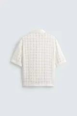 Camisa negra de corte relajado con tejido semitransparente de estructura abierta y patrón a cuadros. Presenta cuello solapa, manga corta, bolsillo plastrón en el pecho y cierre frontal de botones.