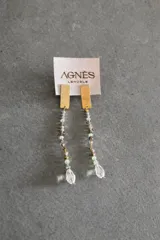 Aros colgantes con base rectangular dorada y tiras de cuentas en tonos beige, gris y verde claro, finalizando con un dije de cristal en forma de gota.