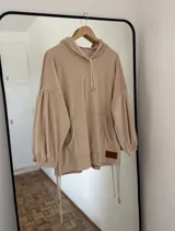 Canguro color beige de plush con capucha, bolsillo frontal y puños fruncidos. Se caracteriza por sus mangas globo y los tajos laterales con tiras ajustables.