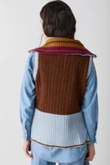 Chaleco de lana con cierre frontal completo y cuello alto. Presenta un diseño de bloques de color en tonos marrón chocolate y celeste, combinando secciones de tejido trenzado con secciones de tejido acanalado. Posee un corte asimétrico, siendo más largo en la parte posterior, y aberturas laterales en el ruedo.