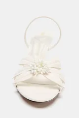 Sandalia blanca de tacón fino con detalle de flor en la parte delantera y tira trasera con elástico ajustable.