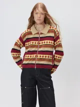 Campera de punto estampada de corte holgado, con cierre metálico frontal y cuello de corderito sintético en color tostado. Presenta un diseño de franjas horizontales con patrones geométricos en tonos bordó, mostaza, marrón y beige.