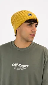 Gorro de lana tejido en punto acanalado color mostaza, con parche circular con logo en la parte frontal.