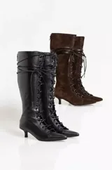 Bota alta de gamuza color marrón oscuro, con diseño de cordones frontales y cierre lateral. Presenta una silueta estilizada con punta fina y un taco bajo tipo kitten heel.