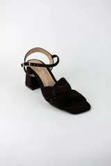 Sandalia de tacón cuadrado medio, confeccionada en material textil tipo gamuza color negro. Presenta un diseño de tiras cruzadas con nudo en la parte delantera y una tira ajustable con hebilla metálica en el tobillo.