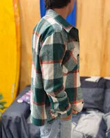 Sobrecamisa de paño con diseño a cuadros en tonos verde, blanco y detalles en naranja. Presenta cuello clásico, cierre frontal con botones, mangas largas con puños abotonados y dos bolsillos frontales con solapa en el pecho.