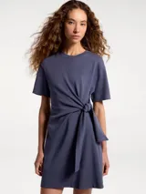 Vestido corto de cuello redondo y manga corta, confeccionado en tejido suave. Presenta un detalle de nudo lateral con lazo ajustable en la cintura que crea un efecto drapeado.