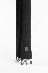 Pashmina de textura plisada en color beige, con flecos en el extremo inferior y un pequeño aplique de cuero sintético con logo grabado.
