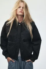 Campera bomber negra, acolchada con patrón de rombos. Presenta cuello, puños y bajo acanalados. Tiene cierre frontal con botones a presión y dos bolsillos de parche en la parte delantera.
