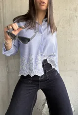 Camisa celeste a rayas verticales blancas, con cuello clásico, cierre frontal con botones y mangas largas. Presenta detalle de bordado calado con motivos florales en el ruedo.