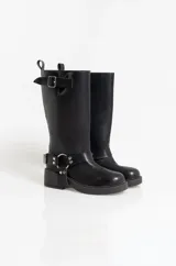 Bota de caña alta estilo biker, confeccionada en cuero sintético negro. Presenta una hebilla ajustable en la parte superior de la caña y un arnés decorativo en el tobillo con argolla metálica y remaches.