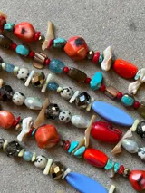 Collar artesanal compuesto por una combinación de cuentas de cristal facetado en tonos gris y bronce, piedras tipo dalmata, cuentas cilíndricas de color crema y cuentas de vidrio azul claro. Presenta un dije central alargado de color azul pastel y terminaciones en cadena de acero.