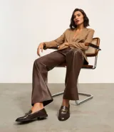 Mocasines de mujer confeccionados en poliuretano color marrón oscuro, con acabado brillante y detalle decorativo metálico dorado en el empeine. Presentan una silueta clásica de punta redondeada y suela plana con pequeño tacón.
