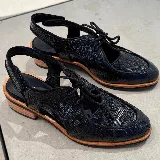 Zapato tipo sueco de cuero vacuno negro con textura reptil, con cordones y talón descubierto. Tiene suela de madera con altura de 4,5 cm en el talón y 1,5 cm en la parte delantera.