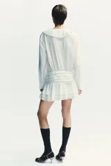 Vestido corto blanco de escote en V y manga larga con puños abotonados. Presenta volantes con bordados de encaje en el cuello, escote y falda. Cierre lateral con cremallera oculta.