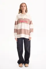 Buzo deportivo sin friza a rayas anchas en tonos blanco y tostado, con cuello polo, escote en V y estampa frontal con la inscripción "California est.1969" en color fucsia. Presenta un corte semi oversize y mangas largas.