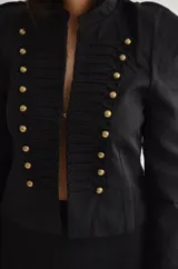 Chaqueta de estilo militar en color negro, con cuello mao y cierre frontal con ganchos decorativos y botones dorados. Presenta hombreras estructuradas y diseño entallado.