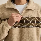 Buzo de polar tipo sherpa en color beige, con cierre frontal parcial y cuello alto. Presenta una banda horizontal estampada con diseño geométrico étnico en tonos marrones y blancos a la altura del pecho.